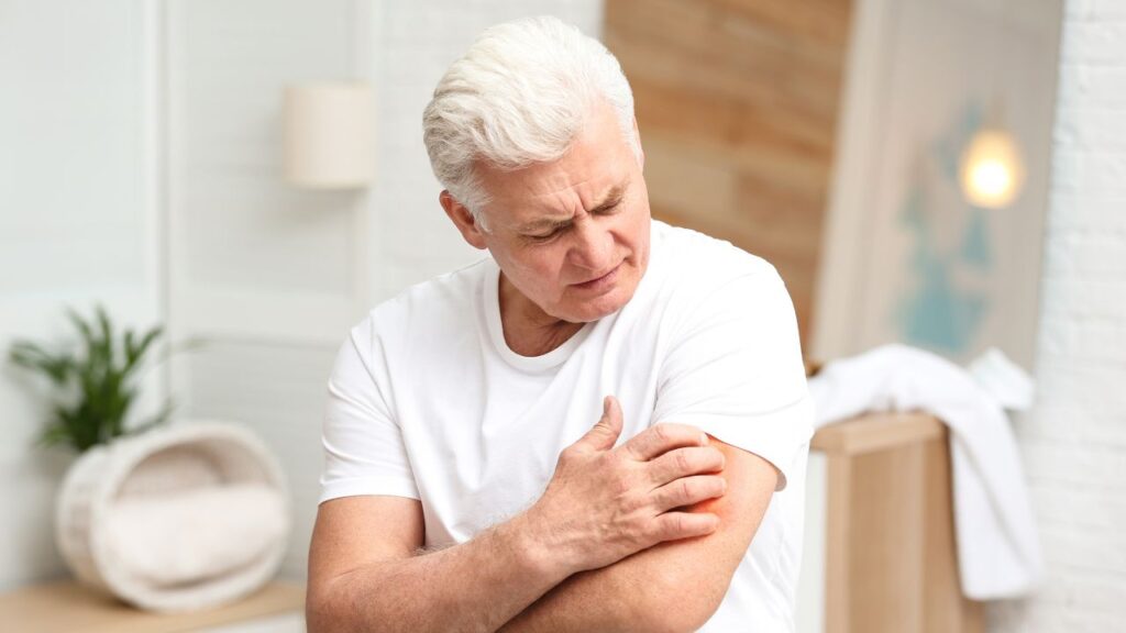 Senior Man Scratching Arm Indoors. Allergy Symptom